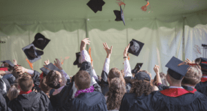 A group of graduates dressed in black caps and gowns celebrate by tossing their mortarboards into the air. The scene takes place under a large tent, with sunlight highlighting the excitement of the moment. The graduates have their hands raised, cheering as their caps, some with orange tassels, fly above them.